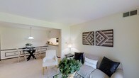 This single-floor residence offers a thoughtfully arranged open-concept layout that seamlessly connects the main living areas. The living room serves as the heart of the home, featuring light neutral walls that create a bright and airy atmosphere. Decorative wooden geometric wall art adds a warm, textural accent to the space, while a glass-top coffee table and a mix of seating — including a plush sofa and a clean-lined white accent chair — provide both comfort and style. A potted indoor plant introduces a touch of greenery, enhancing the welcoming ambiance.

The living area flows naturally into the dining space, which is defined by a pendant light overhead and a glass-top dining table with coordinating chairs, making it an inviting spot for everyday meals. Adjacent to the dining area, the kitchen features a raised breakfast bar countertop in warm wood tones, providing a casual seating or serving area. The kitchen is compact yet functional, with built-in appliances visible along the wall. Throughout the space, light-toned flooring unifies the rooms and contributes to the overall sense of openness and cohesion. The neutral color palette and curated furnishings give the space a modern yet approachable character.