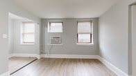 This single-floor unit offers a clean, modern aesthetic throughout, characterized by soft gray walls, crisp white baseboards, and light wood-look luxury vinyl flooring that flows seamlessly through the main living areas. The open layout, as revealed by the floor plan, connects a spacious living area to the kitchen and additional rooms, creating a functional and cohesive flow.

The kitchen is a standout feature, outfitted with bright white shaker-style cabinetry, a classic white subway tile backsplash, and stainless steel appliances including a gas range and full-size refrigerator. A wall-mounted ventilation hood and matte black fixtures add a contemporary edge to the space.

The living area benefits from multiple windows dressed with horizontal blinds, allowing natural light to fill the room. A wall-mounted air conditioning unit provides climate control. An entry area features a solid white door with secure hardware, along with access to a closet.

The bedroom is distinguished by plush wall-to-wall carpeting in a warm neutral tone, offering a softer, quieter retreat. It is finished with the same light gray walls and white baseboards consistent throughout the unit.

The bathroom has been tastefully renovated, featuring floor-to-ceiling white subway tile surrounding a soaking tub and shower, wood-look tile flooring, a white vanity, and coordinating matte black fixtures and hardware, delivering a polished and modern finish.