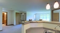 This single-floor residential space is designed with an open-concept layout that seamlessly connects the kitchen and living areas, creating a cohesive and functional living environment. The kitchen features a peninsula-style breakfast bar with an extended countertop that provides casual seating for two, making it ideal for dining or entertaining. A pendant light fixture overhead adds a warm, focused glow to the space. The countertops are finished in a neutral tone that complements the light-colored walls throughout.

The living area transitions naturally from the kitchen, finished with wall-to-wall carpeting that adds comfort underfoot. A large window floods the space with natural light and frames a view of the surrounding greenery, enhancing the sense of openness and connection to the outdoors. A ceiling fan is installed in the adjacent room, providing both air circulation and additional lighting.

Warm wood-toned doors are visible throughout, contributing a classic touch to the otherwise neutral and clean interior palette. The soft, light blue-grey walls give the space a calm and welcoming ambiance. The overall design is practical and comfortable, making efficient use of the single-floor layout while maintaining a bright and airy atmosphere throughout.
