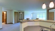 This single-floor residential space is designed with an open-concept layout that seamlessly connects the kitchen and living areas, creating a cohesive and functional living environment. The kitchen features a peninsula-style breakfast bar with an extended countertop that provides casual seating for two, making it ideal for dining or entertaining. A pendant light fixture overhead adds a warm, focused glow to the space. The countertops are finished in a neutral tone that complements the light-colored walls throughout.

The living area transitions naturally from the kitchen, finished with wall-to-wall carpeting that adds comfort underfoot. A large window floods the space with natural light and frames a view of the surrounding greenery, enhancing the sense of openness and connection to the outdoors. A ceiling fan is installed in the adjacent room, providing both air circulation and additional lighting.

Warm wood-toned doors are visible throughout, contributing a classic touch to the otherwise neutral and clean interior palette. The soft, light blue-grey walls give the space a calm and welcoming ambiance. The overall design is practical and comfortable, making efficient use of the single-floor layout while maintaining a bright and airy atmosphere throughout.