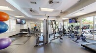 Located at 8800 Pine Forest Rd in Pensacola, FL, this fitness center offers a well-appointed exercise space designed to support a full range of workout routines. The room features a tray ceiling with multiple ceiling fans and recessed skylight panels, keeping the environment well-ventilated and bright. Large floor-to-ceiling windows and sliding glass doors line the walls, flooding the interior with natural light and offering views of the surrounding greenery and outdoor areas.

The exercise floor is covered with durable rubber matting, providing a safe and functional surface throughout. Strength training is well-represented with a multi-station cable machine, a rack of dumbbells, kettlebells, and a dedicated free weights and stretching area complete with exercise mats, stability balls, resistance bands, and a weight bench. Cardio equipment includes elliptical trainers, stationary bikes, treadmills, and a rowing machine, all positioned to take advantage of the natural light and outdoor views.

Wall-mounted flat-screen televisions are positioned throughout the space for entertainment during workouts. Motivational wall decals add an energetic atmosphere to the clean, neutral décor. Full-length mirrors along one wall enhance the sense of openness and allow users to monitor their form. The space is thoughtfully organized to accommodate both group and individual training needs.