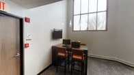 This single-floor commercial space is thoughtfully designed to accommodate both focused work and relaxed seating. The business center area features a dark wood-toned bar-height table paired with warm burnt-orange upholstered stools, providing a functional workspace equipped with desktop computers, a printer, and a wall-mounted flat-screen display. Pendant lighting with sleek, elongated fixtures hangs above the workstation, complementing a large multi-pane window that allows generous natural light to filter in, offering views of mature trees outside.

The adjacent lounge area presents a welcoming atmosphere with grey upholstered armchairs accented by rust-toned throw pillows, arranged around a compact round white side table. The neutral-toned walls are decorated with a set of reclaimed wood-style wall panels, adding a rustic yet contemporary design element. A textured ceiling and patterned carpet flooring contribute to the space's understated, professional character. A wood-paneled door with exit signage provides clear access and reinforces the practical, functional nature of the environment. Overall, the space balances utility with a modern, inviting aesthetic.