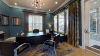 This single-floor professional office space presents a cohesive and polished aesthetic throughout, defined by deep teal walls, crisp white crown molding, and warm tile flooring. The executive office is a standout area, anchored by a large dark desk and complemented by leather seating, a vibrant abstract area rug, and a striking sputnik-style chandelier overhead. Multiple windows dressed with wooden blinds and plaid drape panels fill the room with natural light while offering views of the surrounding exterior. A reception and transitional hallway area connects the spaces seamlessly, featuring a dark console cabinet topped with decorative lamps and framed artwork adorning the walls. This welcoming entry zone provides a professional first impression with ample wall-mounted controls and display elements. Adjacent to the main office, a casual meeting nook offers round bistro-style tables and chairs, ideal for informal gatherings or client consultations. The same sputnik chandelier motif continues here, reinforcing design continuity. Natural light pours in through generously sized windows, and the bold area rug ties the color palette together throughout. The overall ambiance is both functional and visually engaging, blending contemporary design elements with a warm, inviting atmosphere suited for a professional office environment.