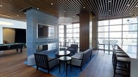 This refined single-floor space exudes contemporary elegance through a carefully curated blend of materials and design elements. The most striking architectural feature is the wood-slat ceiling, which adds warmth and texture while contrasting beautifully with the cooler tones found throughout the room. Recessed lighting integrated into the slats provides a subtle, ambient glow that enhances the sophisticated atmosphere.

The lounge area is anchored by a sleek, modern fireplace set into a smooth concrete-finished accent wall, above which a wall-mounted flat-screen television is prominently displayed. The seating arrangement — consisting of structured armchairs and a bench-style sofa in neutral gray tones — is thoughtfully organized around a pair of dark circular coffee tables, all grounded by a bold blue patterned area rug laid over rich hardwood flooring.

Floor-to-ceiling windows line the perimeter, flooding the space with natural light and offering sweeping views of the surrounding urban skyline. A secondary seating or dining area with bar-height chairs is visible near the windows, further enhancing the functional versatility of the space. A large wooden panel element adds architectural depth and visual separation between zones. The overall ambiance is polished, modern, and well-suited for professional or upscale hospitality use.