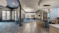 Located at 1 Nathan Pratt Drive in Concord, MA, this single-floor community clubhouse offers a thoughtfully designed gathering space that blends contemporary style with welcoming comfort. The expansive main lounge serves as the heart of the facility, featuring soaring vaulted ceilings, recessed lighting, and a striking combination of large-format polished tile and patterned blue-toned carpeting that defines distinct activity zones. A floor-to-ceiling stacked stone fireplace anchors one end of the lounge, flanked by curated artwork and modern seating arrangements ideal for socializing or relaxing. Adjacent to the fireplace wall, a textured geometric accent panel adds an artistic architectural detail. A sleek billiards table positioned beneath elegant pendant chandeliers provides a dedicated recreation area, while high-top bar seating and café-style tables offer additional gathering spots. The fully equipped kitchen features light wood cabinetry, white quartz countertops, a dark tile backsplash, stainless steel appliances, and a wall-mounted television, making it well-suited for catered events or casual use. A curved lounge nook at one end of the building is enclosed by tall multi-pane windows that flood the space with natural light and offer views of the surrounding neighborhood, creating a quiet retreat within the broader communal environment.