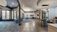 Located at 1 Nathan Pratt Drive in Concord, MA, this single-floor community clubhouse offers a thoughtfully designed gathering space that blends contemporary style with welcoming comfort. The expansive main lounge serves as the heart of the facility, featuring soaring vaulted ceilings, recessed lighting, and a striking combination of large-format polished tile and patterned blue-toned carpeting that defines distinct activity zones. A floor-to-ceiling stacked stone fireplace anchors one end of the lounge, flanked by curated artwork and modern seating arrangements ideal for socializing or relaxing. Adjacent to the fireplace wall, a textured geometric accent panel adds an artistic architectural detail. A sleek billiards table positioned beneath elegant pendant chandeliers provides a dedicated recreation area, while high-top bar seating and café-style tables offer additional gathering spots. The fully equipped kitchen features light wood cabinetry, white quartz countertops, a dark tile backsplash, stainless steel appliances, and a wall-mounted television, making it well-suited for catered events or casual use. A curved lounge nook at one end of the building is enclosed by tall multi-pane windows that flood the space with natural light and offer views of the surrounding neighborhood, creating a quiet retreat within the broader communal environment.