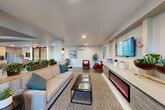 Located at 171 Siesta Way in Sonoma, California, this single-floor community space offers a thoughtfully designed layout that blends comfort and functionality. Wood-plank tile flooring flows throughout, unifying the open-concept interior and lending a warm, modern aesthetic.

The main lounge area is a welcoming focal point, featuring a sleek electric fireplace built into a contemporary media console, a large wall-mounted TV, and comfortable seating including a neutral sofa with teal accent pillows and bold red armchairs. Recessed lighting and a wall-mounted mini-split system ensure year-round comfort, while artwork and curated décor add a polished, residential feel.

Adjacent to the lounge, a spacious dining area accommodates multiple guests with a large dark-wood table and mixed seating. The kitchen behind it is well-equipped with light wood cabinetry, a patterned tile backsplash, stainless steel refrigerator, microwave, and generous open shelving for storage.

A corridor with built-in cabinetry connects the main areas to a quieter seating nook featuring sheer-draped windows that invite natural light and outdoor views. Large glass-panel entry doors further brighten the space and provide a visual connection to the exterior. A dedicated office rounds out the layout, offering a private area for focused work or meetings.
