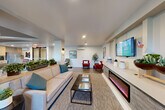 Located at 171 Siesta Way in Sonoma, California, this single-floor community space offers a thoughtfully designed layout that blends comfort and functionality. Wood-plank tile flooring flows throughout, unifying the open-concept interior and lending a warm, modern aesthetic.

The main lounge area is a welcoming focal point, featuring a sleek electric fireplace built into a contemporary media console, a large wall-mounted TV, and comfortable seating including a neutral sofa with teal accent pillows and bold red armchairs. Recessed lighting and a wall-mounted mini-split system ensure year-round comfort, while artwork and curated décor add a polished, residential feel.

Adjacent to the lounge, a spacious dining area accommodates multiple guests with a large dark-wood table and mixed seating. The kitchen behind it is well-equipped with light wood cabinetry, a patterned tile backsplash, stainless steel refrigerator, microwave, and generous open shelving for storage.

A corridor with built-in cabinetry connects the main areas to a quieter seating nook featuring sheer-draped windows that invite natural light and outdoor views. Large glass-panel entry doors further brighten the space and provide a visual connection to the exterior. A dedicated office rounds out the layout, offering a private area for focused work or meetings.