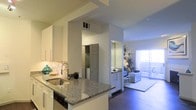This single-floor residence offers a thoughtfully designed open-concept layout that promotes a seamless flow between living spaces. The modern kitchen is a standout feature, showcasing granite countertops, white shaker-style cabinetry, a stainless steel sink, and a stylish mosaic tile backsplash. A breakfast bar extends from the counter, providing a casual dining or entertaining space. Built-in appliances, including a dishwasher, are neatly integrated into the cabinetry for a clean, streamlined look. From the kitchen, sightlines extend directly into the living area, which is finished with rich hardwood-style flooring and benefits from generous natural light streaming through large, floor-to-ceiling windows or sliding glass doors. The bright, airy living space is anchored by a plush area rug and opens to what appears to be a balcony or patio beyond the vertical blinds. A utility or laundry area with stacked units is conveniently tucked behind a door just off the kitchen. The corridor leading to additional spaces features consistent cabinetry and a mirror, reinforcing the cohesive, contemporary aesthetic found throughout. The overall ambiance is bright, clean, and functional.