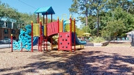 Located at 1551 Sam Rittenberg Blvd in Charleston, South Carolina, this outdoor recreational space offers a well-equipped playground set within a natural, tree-lined environment. The centerpiece of the area is a vibrant, multi-component play structure finished in bold red, yellow, and blue tones. It features a rock climbing wall, multiple slides, elevated platforms, and interconnected activity panels, providing a variety of engaging options for active play. The ground beneath the structure is covered with a wood chip surface, offering a natural and protective layer throughout the play zone. Tall pine and deciduous trees surround the perimeter, casting dappled shade across the grounds and creating a pleasant, shaded atmosphere on sunny days. The space is enclosed by a sturdy chain-link fence along its borders, which backs up to a dense treeline, offering both security and a sense of seclusion from the surrounding area. A glimpse of a pool with yellow shade umbrellas is visible in the background, suggesting additional recreational amenities nearby. The open layout of the grounds provides ample room for free play beyond the main structure, making this a well-rounded outdoor destination for active recreation.