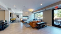 This well-appointed single-floor amenity space showcases a contemporary design aesthetic with light hardwood flooring, neutral tones, and curated furnishings throughout. The expansive main lounge serves as the centerpiece, featuring a stunning floor-to-ceiling marble-clad fireplace wall, a comfortable leather sectional, and mid-century modern accent chairs. Large sliding glass doors flood the space with natural light and provide direct access to an outdoor terrace with seating.

A secondary lounge area offers additional seating with a wall-mounted TV, open shelving, and a cozy reading nook, while sliding barn doors connect seamlessly to a dedicated dining room anchored by a bold chandelier and a large communal table. A smaller, intimate dining nook with a round glass table and a striking gold ring pendant light offers an alternative gathering option with outdoor views.

The full-service kitchen is a standout feature, boasting a dramatic geometric metallic tile backsplash, blue-grey cabinetry, stainless steel appliances, a large island with bar seating, and elegant gold geometric pendant lights. A billiards area rounds out the space, providing a recreational retreat. Throughout, modern artwork, purposeful lighting, and consistent finishes create a cohesive and inviting communal environment.