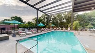 Located at 710 Greenville St in LaGrange, Georgia, this outdoor pool facility offers a well-appointed aquatic retreat designed for relaxation and leisure. The centerpiece is a generously sized swimming pool with crystal-clear turquoise water, bordered by a spacious concrete pool deck that provides ample room for sunbathing and socializing.

A striking architectural feature is the partially covered steel pergola structure that extends over a portion of the deck, offering shade while maintaining an open-air feel with views of the surrounding sky. The covered section, finished with a corrugated metal roof, shelters multiple seating arrangements, including wicker egg chairs and sectional sofas with vibrant orange cushions, creating comfortable lounging zones.

Along the sun-drenched side of the pool, a row of lounge chairs paired with teal umbrellas invites guests to unwind in the open air. A notable decorative element is a sculptural figure positioned near the covered area, adding a unique character to the space. The perimeter is enclosed by black iron fencing, with mature trees providing a lush, green backdrop and a sense of natural privacy. The combination of covered and open-air spaces, decorative touches, and resort-style furnishings makes this pool area a versatile and inviting outdoor destination.