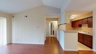 This well-appointed single-floor residence offers a thoughtfully designed layout with a warm, neutral palette throughout. The living area is an immediate standout, featuring dramatic vaulted ceilings that amplify the sense of space and an abundance of natural light streaming through a large sliding glass door fitted with vertical blinds. A classic fireplace with a tiled surround anchors one wall, adding a cozy focal point to the room. Rich, warm-toned hardwood flooring flows seamlessly across the living, dining, and bedroom areas, lending the home a cohesive and inviting character.

The galley-style kitchen is efficiently designed with ample rich wood cabinetry, light countertops, a full suite of white appliances including a range and refrigerator, an overhead range hood, and a double sink. Track lighting illuminates the workspace, while light tile flooring completes the functional aesthetic. A breakfast bar peninsula connects the kitchen to the main living area, providing a convenient casual dining option.

The bedroom is generously proportioned with continued hardwood flooring, a ceiling fan with light fixture, and a walk-in closet offering practical storage. The full bathroom features rich wood vanity cabinetry, a single-basin sink with light countertops, a tiled tub-shower combination, and light tile flooring throughout. A hallway connects the primary spaces, with additional closet storage visible off the main corridor.