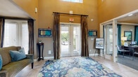 This impressive communal space makes a striking first impression with a welcoming entryway framed by floor-to-ceiling glass double doors, draped in rich bronze silk curtains that complement the warm golden walls. The foyer opens into a spacious reception area finished in large-format neutral tile, where structural columns define distinct functional zones, including a refreshment station and an information desk, each accented with stylish pendant lighting.

The main lounge area commands attention with dramatic double-height ceilings and an expansive wall of windows dressed in matching bronze drapery, flooding the interior with abundant natural light. Slate-blue walls provide a refined backdrop for plush leather sofas, glass-topped coffee tables, and vibrant floral-patterned area rugs in blue and gold tones that anchor each seating arrangement.

The focal point of the lounge is a towering floor-to-ceiling natural stone fireplace, topped by a mounted flat-screen television and flanked by second-floor balconies with wrought iron railings. Wall sconces cast a warm, ambient glow throughout. Additional seating alcoves with accent lighting, tropical greenery, and curated artwork contribute to an atmosphere that is both polished and inviting, offering a comfortable, resort-style communal environment.