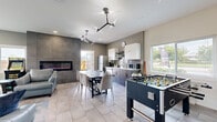 Located at 1459 KLO Road in Kelowna, BC, this single-floor common area offers a thoughtfully designed open-concept layout that seamlessly blends a fully equipped kitchen, dining area, and living space. The interior is finished with large-format light tile flooring throughout, complemented by contemporary grey walls and crisp white ceilings with modern branching light fixtures that add a striking architectural touch.

The kitchen features two-tone cabinetry — dark lower cabinets with white uppers — a grey subway tile backsplash, a full-size stainless steel refrigerator, microwave, and ample counter space. A large dining table with upholstered chairs sits centrally, ideal for group gatherings. A foosball table adds a recreational focal point to the space.

The living area is bright and welcoming, with large windows allowing generous natural light and views of the surrounding greenery. Comfortable leather seating, a flat-screen TV, and a classic arcade game cabinet make this a versatile entertainment zone. A welcoming entry nook features plush blue accent chairs, wall-mounted board games, and lush indoor plants. A floor-to-ceiling tiled accent wall with a sleek linear fireplace anchors the space with warmth and modern elegance.