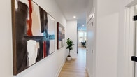 This 1-bedroom, 1-bathroom unit is laid out across a single floor, making efficient use of its space with a well-considered floor plan. Light wood-tone flooring runs throughout, creating a cohesive and airy aesthetic that connects each room seamlessly.

The entry hallway sets an inviting tone, featuring crisp white walls adorned with bold abstract artwork and recessed lighting overhead. A potted fiddle-leaf fig plant adds a natural accent, while the corridor opens directly into the living area and kitchen beyond.

The living room is bright and comfortably scaled, with clean white walls and light carpeting. It features a stylish arrangement including a green velvet sofa, a round marble-topped coffee table, and a distinctive gold organic-form mirror, complemented by framed artwork and warm lamp lighting.

The kitchen, visible from the living area, is outfitted with dark cabinetry, stainless steel appliances including a refrigerator and range, and a functional layout suited for everyday cooking.

The bedroom offers a cozy retreat with a ceiling fan, a striking black-hardware barn door, and clean white walls decorated with framed prints. A dedicated closet provides practical storage.

The bathroom is finished in a modern monochromatic palette, featuring floor-to-ceiling white subway tile surrounding the tub-shower combo, matte black fixtures and hardware, a floating dark vanity with a white countertop, and light wood flooring—delivering a sharp, contemporary look.