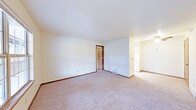 This single-floor space features a spacious main living area finished with wall-to-wall carpet in a neutral tone, providing a comfortable and practical foundation throughout. The room is characterized by clean white walls, dark wood baseboards, and a textured ceiling that adds subtle visual interest overhead. A floor vent indicates a forced-air system integrated into the flooring. The open layout connects seamlessly to an adjoining area visible to the right, which appears to include additional built-in or appliance features, suggesting a utility or kitchen space. A doorway with a dark wood-framed door leads to a secondary room, likely a bedroom or storage area, offering clear spatial separation between living zones. The overall design is straightforward and functional, with a neutral color palette throughout that allows for versatile furnishing options. The layout maximizes usable floor space, making the area feel open and adaptable. The consistent flooring and wall finishes throughout create a cohesive, uniform aesthetic suited to practical, everyday living.