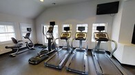 This fitness center is housed in a single-floor space featuring a clean, functional design with white walls and vaulted ceilings that contribute to an open, airy atmosphere. The room is well-lit by natural light streaming through multiple windows along the exterior wall, each framed with decorative iron grating that adds a subtle architectural detail. A glass-panel door provides additional light and outdoor access.

The cardio equipment lineup includes three treadmills and at least two elliptical machines, each unit equipped with individual digital display screens for personalized workout tracking. A wall-mounted flat-screen television is positioned at an elevated angle for viewing during exercise sessions. The flooring is a durable, textured dark surface suited for high-traffic fitness use, while recessed lighting supplements natural light for consistent illumination throughout the room. The neutral color palette and uncluttered layout ensure the focus remains on functionality, making this space a practical and well-appointed facility for cardiovascular training.