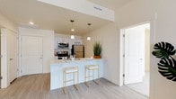 This single-floor residence is designed with a clean, contemporary aesthetic characterized by warm neutral tones, light-toned luxury vinyl plank flooring in the main living areas, and plush carpet in the bedrooms. The open-concept kitchen serves as a central hub, anchored by a white peninsula with an integrated sink and seating for two. Stainless steel appliances—including a refrigerator, range, microwave, and dishwasher—are paired with white cabinetry and light countertops, creating a bright, functional workspace. Pendant lighting above the peninsula adds a modern design touch. The kitchen flows seamlessly into the living area, which offers an open, airy feel with access to additional rooms. The bedrooms are comfortably sized, each featuring neutral carpet flooring, white walls, and windows that allow natural light and views of the surrounding neighborhood. Two full bathrooms are finished in a consistent white palette, each equipped with a tub-shower combination, a single-sink vanity with white cabinetry, and tile flooring. The overall ambiance throughout the unit is bright, clean, and move-in ready, with a cohesive modern design sensibility.