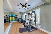 Located on Zierdt Road SW in Huntsville, Alabama, this fitness center offers a thoughtfully designed exercise space across a single open floor. The room is finished with soft sage-green walls, crisp white crown molding, and light hardwood flooring that lend a clean, welcoming ambiance throughout. Large windows fitted with white plantation shutters allow abundant natural light to fill the space, while multiple large-blade ceiling fans ensure comfortable airflow during workouts.

The cardio zone features a treadmill, elliptical trainer, stationary bike, and rowing machine, each placed on protective floor mats and positioned to take advantage of the natural light and wall-mounted flat-screen television. A dedicated strength training area houses a heavy-duty power rack with a weight bench and an assortment of Olympic plates, alongside a cable machine for versatile resistance training. Floor-to-ceiling mirrors line one wall, visually expanding the space and aiding in form checks during exercise.

Organizational racks display a colorful array of medicine balls and stability balls, while a separate open area provides ample room for stretching, yoga, or functional training. A built-in water fountain station with dual dispensers and a convenient waste receptacle are positioned near the glass exterior door, which opens directly to the outdoors. Recessed lighting complements the natural illumination, completing this polished and fully appointed fitness facility.