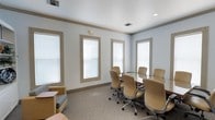 This single-floor conference room offers a clean, professional environment well-suited for meetings and collaborative work. The room is anchored by a rectangular conference table finished in a dark tone, surrounded by several tan upholstered ergonomic chairs that provide comfortable seating for attendees. The layout allows for easy movement around the table, giving the room a functional yet open feel.

The walls are painted in a soft, neutral light blue-gray, complemented by warm beige trim and crown molding that adds a subtle architectural detail. Three large windows fitted with white horizontal blinds line the walls, allowing for generous natural light while offering privacy and glare control. Recessed ceiling lighting supplements the natural light, ensuring consistent illumination throughout the space.

The floor is covered in a neutral, low-pile carpet with a subtle pattern, contributing to the room's understated and professional aesthetic. A small seating area with a compact sofa and side table is visible in one corner, providing a secondary informal space within the room. Overall, the space presents a tidy, well-organized atmosphere appropriate for business use.