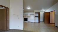 This single-floor space features an open-concept design that connects the living area to the kitchen, creating a cohesive and functional layout. The kitchen is equipped with white cabinetry providing ample storage, a black electric range with an over-the-range microwave, a white refrigerator, and a dishwasher. The flooring in the kitchen and adjoining areas is a neutral-toned vinyl, while the living space features carpet, offering a comfortable transition between zones.

A notable feature is the in-unit laundry area, which includes a stacked washer and dryer unit neatly positioned in a dedicated alcove with wood-toned cabinetry. This practical addition adds significant convenience. The ceilings are of standard height throughout, and flush-mount light fixtures provide consistent overhead lighting across the space.

Neutral wall tones in soft white and beige contribute to a clean, understated aesthetic. Multiple doorways are visible, suggesting well-distributed room access. A structural column divides the open living and kitchen areas, adding a subtle architectural element. Overall, the space offers a practical and straightforward layout suited for everyday living.