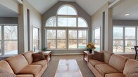 Located in Fort Mitchell, Kentucky, this single-floor community space is defined by its striking architectural character and welcoming atmosphere. The centerpiece of the space is a grand sunlit lounge featuring soaring vaulted ceilings and a dramatic floor-to-ceiling window wall crowned by a sweeping arched transom. This impressive glazing fills the room with abundant natural light while framing picturesque views of the surrounding wooded landscape and an outdoor deck with a pool area beyond. Two comfortable sofas flank a stone-topped coffee table on plush carpeting, creating an inviting gathering area.

A long central corridor connects the main lounge to the rest of the space, with vaulted ceilings continuing overhead and a ceiling fan providing comfort. Multiple doors line the hallway, offering access to additional rooms and a glazed entryway that opens to the outdoors. A secondary lounge area features built-in cabinetry with shelving, a television, and French doors leading to the deck, providing a relaxed media and seating retreat.

The kitchen and dining area are thoughtfully designed with warm hardwood flooring, a peninsula breakfast bar with bar-height seating, light wood cabinetry, and a full suite of appliances. An adjacent casual dining nook with a round pub-height table enjoys direct deck access through a sliding door, keeping the connection to the outdoors close at hand throughout the space.