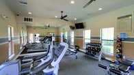 This fitness center is a thoughtfully designed, single-floor facility built to support a full range of workout routines. The space is outfitted with a variety of cardio equipment, including treadmills and elliptical machines, arranged to allow comfortable movement throughout the floor. A dedicated free weight area features a multi-tiered dumbbell rack stocked with a wide selection of weights, accompanied by a flat bench for strength training exercises.

Large windows line the walls, flooding the interior with natural light and providing views of the surrounding greenery, creating an energizing atmosphere for exercise. Wall-mounted mirrors extend along key sections of the room, visually expanding the space and allowing users to monitor their form during workouts. Recessed ceiling lights supplement the natural light, ensuring the facility is well-lit at all times.

Two ceiling fans are mounted overhead, promoting consistent airflow and keeping the environment comfortable during intense sessions. A wall-mounted flat-screen television adds an entertainment element to the space. The flooring features durable, cushioned carpet suited for a fitness environment. Warm neutral tones with a contrasting horizontal accent stripe along the walls contribute to the clean, welcoming aesthetic of this well-appointed fitness facility.