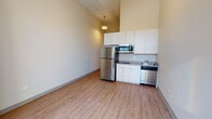 This single-floor unit presents a clean, modern aesthetic throughout its well-organized layout. The living area is characterized by notably tall ceilings that amplify the sense of space, with large windows that flood the interior with natural light while offering views of the surrounding urban skyline. Light wood-tone laminate flooring runs consistently throughout, lending warmth and continuity to each area.

The kitchen is efficiently designed and equipped with a full suite of stainless steel appliances, including a full-size refrigerator, built-in microwave, two-burner electric cooktop, and dishwasher. White cabinetry with neutral countertops provides ample storage and workspace, complemented by a single-basin sink. A pendant light fixture adds a subtle contemporary touch overhead.

A corridor connects the main living spaces, featuring a panel-style sliding door that offers flexible access to adjoining areas. The bathroom is appointed with a tub-shower combination, a wall-mounted vanity with an integrated sink, and a large mirror, all finished in a clean, neutral palette. The overall ambiance of the unit is bright, functional, and move-in ready, reflecting a straightforward modern design sensibility throughout.