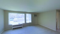 This single-floor space centers around a spacious, open living area finished with neutral beige walls and light-colored carpeting that lends a clean, understated aesthetic throughout. The room is characterized by an abundance of natural light, drawn in through multiple large windows dressed with vertical blinds that allow for flexible light control. The windows offer views of the surrounding outdoor environment, contributing to an airy and open atmosphere.

Practical heating and cooling infrastructure is visibly integrated into the space: baseboard radiator units run beneath the windows along the perimeter, while a wall-mounted air conditioning unit provides additional climate control. The smooth, light-toned ceiling complements the overall neutral palette, maintaining a consistent and cohesive look.

The layout is open and unobstructed, offering versatile potential for furnishing and use. White baseboards frame the carpeted floor cleanly, adding a modest architectural detail. Overall, the space presents a functional and simply designed interior, well-suited for a variety of living arrangements.
