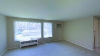 This single-floor space centers around a spacious, open living area finished with neutral beige walls and light-colored carpeting that lends a clean, understated aesthetic throughout. The room is characterized by an abundance of natural light, drawn in through multiple large windows dressed with vertical blinds that allow for flexible light control. The windows offer views of the surrounding outdoor environment, contributing to an airy and open atmosphere.

Practical heating and cooling infrastructure is visibly integrated into the space: baseboard radiator units run beneath the windows along the perimeter, while a wall-mounted air conditioning unit provides additional climate control. The smooth, light-toned ceiling complements the overall neutral palette, maintaining a consistent and cohesive look.

The layout is open and unobstructed, offering versatile potential for furnishing and use. White baseboards frame the carpeted floor cleanly, adding a modest architectural detail. Overall, the space presents a functional and simply designed interior, well-suited for a variety of living arrangements.