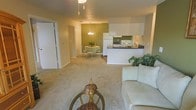 This single-floor residence showcases an open-concept design that seamlessly integrates the living, dining, and kitchen areas into one cohesive, functional space. The living area is anchored by plush carpeting in a neutral beige tone, providing a soft and comfortable foundation. A cozy sofa and a decorative wicker accent table furnish the space, complemented by potted greenery that adds a natural touch.

The kitchen features a peninsula-style counter with dark countertops and white cabinetry, offering both workspace and a visual divider between the cooking and living zones. White appliances, including a refrigerator and stove, contribute to a clean, uniform aesthetic. A pass-through opening above the counter enhances the sense of connectivity throughout the space.

Adjacent to the kitchen, the dining area is distinguished by a bold sage green accent wall that adds depth and character. A chandelier with a classic design hangs above a small round dining table surrounded by chairs, creating an intimate setting. Consistent carpeting flows throughout, unifying each zone, while the textured ceiling and recessed lighting complete the understated, functional ambiance of the interior.