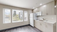 This efficiently designed single-floor unit offers a clean, modern aesthetic with a neutral color palette and wood-look flooring that flows seamlessly throughout the space. The kitchen is a standout feature, outfitted with light wood-grain cabinetry that extends to the ceiling for maximum storage, a white refrigerator, microwave, and a single-basin sink with modern fixtures. A large window fills the kitchen and main living area with natural light, offering views of surrounding greenery and neighboring structures. A wall-mounted panel radiator provides efficient heating to the open living area, keeping the space comfortable year-round. The unit includes a generously sized closet area with dual wire shelving rods, offering practical and organized storage. The full bathroom is finished with large-format marble-look tile surrounding the soaking tub and shower, complemented by wood-look tile flooring, a toilet, and a wall-mounted sink. Recessed lighting in the kitchen adds a polished touch, while the overall design prioritizes functionality and clean lines, making this a well-appointed and practical living space.