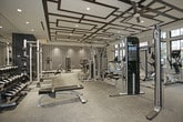 This fitness center is a thoughtfully designed, single-floor facility that seamlessly blends high-end aesthetics with comprehensive workout functionality. The space is divided into distinct training zones, each purposefully outfitted to support a full range of exercise disciplines.

The cardio zone is positioned along a wall of large, multi-pane black-framed windows that flood the area with natural light and provide views of the surrounding outdoor environment. Treadmills, stationary bikes, and elliptical machines are arranged to take advantage of the outlook, and wall-mounted televisions enhance the experience.

An adjacent functional training area features a cable crossover machine and additional cardio equipment, set beneath a striking coffered ceiling with an oversized ceiling fan. Full-length mirrored walls visually expand the space, while a dedicated spin studio enclosed in glass walls offers an immersive cycling experience with its own mounted display screen.

The strength training zone is anchored by a full power rack, a well-stocked dumbbell rack, and a flat bench, all set against deep navy accent walls and mirrored surfaces. A separate selectorized machine area is set against a dramatic geometric-patterned wallcovering in navy and gold, illuminated by recessed accent lighting that adds a sophisticated, boutique gym ambiance throughout. Commercial-grade carpet flooring ties all zones together cohesively.