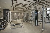 This fitness center is a thoughtfully designed, single-floor facility that seamlessly blends high-end aesthetics with comprehensive workout functionality. The space is divided into distinct training zones, each purposefully outfitted to support a full range of exercise disciplines.

The cardio zone is positioned along a wall of large, multi-pane black-framed windows that flood the area with natural light and provide views of the surrounding outdoor environment. Treadmills, stationary bikes, and elliptical machines are arranged to take advantage of the outlook, and wall-mounted televisions enhance the experience.

An adjacent functional training area features a cable crossover machine and additional cardio equipment, set beneath a striking coffered ceiling with an oversized ceiling fan. Full-length mirrored walls visually expand the space, while a dedicated spin studio enclosed in glass walls offers an immersive cycling experience with its own mounted display screen.

The strength training zone is anchored by a full power rack, a well-stocked dumbbell rack, and a flat bench, all set against deep navy accent walls and mirrored surfaces. A separate selectorized machine area is set against a dramatic geometric-patterned wallcovering in navy and gold, illuminated by recessed accent lighting that adds a sophisticated, boutique gym ambiance throughout. Commercial-grade carpet flooring ties all zones together cohesively.