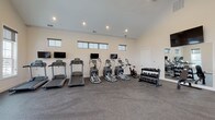 Located at 3986 Parkmead Dr in Grove City, Ohio, this fitness center is a thoughtfully designed, single-floor facility built to support a well-rounded workout experience. The space features soaring vaulted ceilings that create an open, airy atmosphere, complemented by rows of clerestory windows along the upper walls that flood the interior with natural light. Durable rubber flooring throughout provides comfort and safety during exercise. The cardio zone is well-appointed with multiple treadmills and elliptical machines, each paired with its own flat-screen monitor, along with a stationary spin bike. A large wall-mounted mirror expands the sense of space while supporting form-focused training. For strength training, the facility offers a multi-function cable and weight machine, a full rack of dumbbells in varying weights, and additional free-weight accessories. A dedicated open area provides ample room for stretching, functional movement, or floor exercises. Near the entrance, a water station offers convenient hydration access alongside posted workout reference materials. Glass-paneled exterior doors connect the space to the outdoors, adding to the bright and welcoming environment. The clean, neutral color palette and recessed lighting contribute to a focused and professional fitness atmosphere.