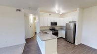 This single-floor residence offers a clean, modern aesthetic throughout, combining functional design with comfortable living spaces. The open-concept kitchen serves as a central highlight, featuring crisp white shaker-style cabinetry, granite countertops, and a full suite of stainless steel appliances including a refrigerator, range, and built-in microwave. A peninsula with a stainless steel sink extends the counter space and creates a natural divide between the kitchen and the adjoining living area. Wood-look flooring runs through the kitchen and entry areas, providing a durable and stylish foundation. The living room is bright and welcoming, anchored by a fireplace with a built-in display niche above it, adding architectural character to the space. Plush carpeting and a sliding glass door that allows natural light complete the living area. The bedrooms are generously proportioned, each finished with soft carpeting, neutral walls, and ample closet access. Natural light filters in through large windows, creating an airy atmosphere. The bathroom is well-equipped with a granite-topped vanity, white cabinetry, a large mirror, and a full tub-shower combination with clean tile surround. Throughout the space, a consistent palette of warm neutrals and white finishes contributes to a cohesive and inviting ambiance.