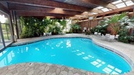 This impressive indoor pool facility offers a resort-like atmosphere defined by thoughtful architectural design and abundant tropical greenery. At its center sits a generously sized freeform swimming pool with vivid turquoise water, framed by a striking exposed wood beam ceiling with skylights that flood the space with natural light throughout the day. The stone-patterned tile deck surrounds the pool with ample room for movement, and is furnished with white lounge chairs, chaise lounges, and casual dining sets, creating comfortable spaces for relaxation and leisure.

Lush tropical plantings — including palm trees, fan palms, elephant ear plants, and a variety of tropical foliage — are thoughtfully integrated throughout the interior, lending the space a verdant, garden-like atmosphere. Warm brick accent walls complement the rich wood ceiling structure, reinforcing a natural, earthy aesthetic. A dedicated hot tub area is nestled among the greenery, offering a more intimate retreat within the larger facility.

Large floor-to-ceiling glass walls and wide glass doors connect the indoor pool seamlessly to a second outdoor pool and sun deck beyond, expanding the recreational options and enhancing the sense of openness. The dual indoor-outdoor layout makes this facility a year-round destination, equally suited for active swimming and quiet relaxation in a lush, sheltered environment.