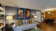 This single-story space showcases a thoughtfully designed living area that balances comfort with visual interest. The living room serves as the focal point, featuring a light gray sofa adorned with vibrant multicolored accent pillows in chevron and floral patterns, adding an energetic contrast to the neutral base. The walls are painted in a cool, deep gray tone, providing a sophisticated backdrop that highlights two large framed floral paintings with bold orange, red, and teal hues, mounted side by side for a striking gallery effect. Crown molding lines the upper edges of the walls, lending a refined architectural detail to the room. Warm hardwood flooring extends throughout, adding texture and continuity. A dark wood side table beside the sofa holds a classic table lamp, contributing soft ambient lighting to the space. An open passageway visible from the living area leads to an adjacent room, suggesting an open and connected floor plan. A glimpse of additional artwork and dining furniture in the neighboring space hints at a cohesive, well-curated interior design aesthetic carried throughout the home.