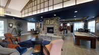 The Avenue presents a thoughtfully designed single-floor space that seamlessly blends rustic charm with contemporary style. At the heart of the main gathering area stands a striking floor-to-ceiling stacked stone fireplace, serving as the room's defining focal point and adding warmth and texture to the space. The open-concept layout connects multiple functional zones, including a comfortable lounge area anchored by a deep navy area rug, upholstered seating, and a round coffee table, creating an inviting setting for relaxation and socializing. A dining area with elegant seating is positioned near natural light-filled windows, while a bar or counter space is visible in the background, enhancing the entertaining potential of the room. The ceiling design is particularly noteworthy, featuring a dramatic two-tone treatment with dark recessed-lit panels contrasted by an exposed, latticed wood beam structure above, adding architectural depth and visual interest. Large tile flooring runs throughout, providing a clean and durable foundation that ties the various zones together. The overall ambiance is sophisticated yet approachable, making it a versatile space suited for both casual gatherings and more formal occasions.