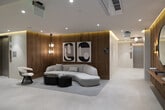 Located at 1655 Malcolm Ave in the Westwood neighborhood of Los Angeles, this single-floor lobby space exemplifies contemporary design with a refined, minimalist aesthetic. The layout flows seamlessly from a sleek, light-toned corridor to a welcoming lounge area, creating a cohesive and polished entry experience.

The corridor is finished in soft, neutral tones with large-format light stone flooring and recessed cove lighting that casts a warm ambient glow along the ceiling line. A built-in wall niche accented with fresh orchid arrangements adds an organic, welcoming touch, while a floating wall-mounted desk with a rich walnut wood backing panel serves as a functional concierge or reception point.

The lounge area is the design centerpiece of the space, featuring floor-to-ceiling vertical wood slat wall paneling that introduces warmth and texture throughout. A sculptural, curved white sofa anchors the seating arrangement, paired with a set of dark, organically shaped coffee tables atop a geometric area rug. Bold black-and-white abstract artwork adorns the walls, complemented by a round mirror and warm wall sconces that enhance the sophisticated atmosphere. A statement pendant chandelier with geometric detailing adds vertical interest and illumination.

Elevator access is integrated discreetly into the space, maintaining the clean architectural lines. Overall, this lobby delivers a high-end, design-forward environment that is both functional and visually impressive.