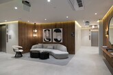Located at 1655 Malcolm Ave in the Westwood neighborhood of Los Angeles, this single-floor lobby space exemplifies contemporary design with a refined, minimalist aesthetic. The layout flows seamlessly from a sleek, light-toned corridor to a welcoming lounge area, creating a cohesive and polished entry experience.

The corridor is finished in soft, neutral tones with large-format light stone flooring and recessed cove lighting that casts a warm ambient glow along the ceiling line. A built-in wall niche accented with fresh orchid arrangements adds an organic, welcoming touch, while a floating wall-mounted desk with a rich walnut wood backing panel serves as a functional concierge or reception point.

The lounge area is the design centerpiece of the space, featuring floor-to-ceiling vertical wood slat wall paneling that introduces warmth and texture throughout. A sculptural, curved white sofa anchors the seating arrangement, paired with a set of dark, organically shaped coffee tables atop a geometric area rug. Bold black-and-white abstract artwork adorns the walls, complemented by a round mirror and warm wall sconces that enhance the sophisticated atmosphere. A statement pendant chandelier with geometric detailing adds vertical interest and illumination.

Elevator access is integrated discreetly into the space, maintaining the clean architectural lines. Overall, this lobby delivers a high-end, design-forward environment that is both functional and visually impressive.