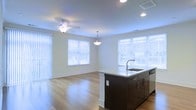 This well-appointed single-floor space offers an open-concept design that seamlessly connects the kitchen and living areas, creating a sense of spaciousness and ease of movement. The kitchen is anchored by a striking dark-finished island with a light stone countertop and an integrated sink, providing ample workspace and a natural gathering point. The island is complemented by a stainless steel dishwasher and clean, modern cabinetry hardware. Recessed lighting and a decorative pendant light above the island ensure the kitchen is well-illuminated throughout the day and evening.

The adjoining living area is bathed in natural light, courtesy of multiple large windows dressed with white horizontal blinds and vertical panel blinds that allow for flexible light control. Light-toned hardwood flooring extends throughout the open space, adding warmth and continuity to the design. A ceiling fan with a light fixture provides both comfort and additional illumination in the living zone. The walls are finished in a crisp, neutral white, lending a clean and versatile backdrop that complements a variety of décor styles. Crown molding adds a refined architectural detail throughout, elevating the overall finish of the space.