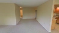 This single-floor space presents a straightforward, functional layout centered around an open main living area. The room features wall-to-wall carpeting in a neutral beige tone that extends throughout, providing a cohesive and comfortable foundation underfoot. The walls are finished in a soft, warm off-white that contributes to the bright and airy feel of the interior, complemented by standard-height ceilings that maintain a practical sense of openness.

From the main living area, two distinct doorways are visible. One opening leads to an adjacent room, likely a bedroom or secondary living space, and appears to include a closet or storage area nearby. A second doorway leads further into the unit, where a ceiling light fixture is visible, suggesting a hallway or additional room with its own lighting. The overall layout implies a connected series of rooms accessible from the central living space.

The unit is currently vacant, allowing for a clear view of the space's clean lines and neutral palette. The minimalist design and lack of ornamentation make it highly adaptable to a variety of furnishing styles and personal preferences, offering a blank canvas for future occupants.