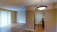 This single-floor residence offers a practical and comfortable layout designed for easy everyday living. The open-concept living area is finished with rich, dark hardwood flooring that extends throughout the space, providing a warm and cohesive aesthetic. Neutral-toned walls in soft beige complement the flooring and create a calm, inviting ambiance. A structural column subtly defines the transition between the living area and the kitchen, maintaining an open feel while offering a natural sense of separation. The living area is generously sized, providing ample room for a variety of furniture arrangements, and features clean white baseboards that add a crisp finishing touch. The kitchen is appointed with dark wood cabinetry, a granite or stone-look countertop, and a black refrigerator, creating a functional and stylish cooking space. A striking circular pendant light with a dark wood frame and frosted glass shade hangs above, serving as a focal point and providing warm overhead illumination. The textured ceiling adds a subtle layer of detail to the interior. Overall, the space presents a clean, well-maintained environment with a consistent design language that balances warmth and functionality throughout.
