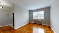 This single-floor unit offers a straightforward, functional layout across several well-defined rooms, all unified by warm-toned hardwood flooring that runs throughout the living areas and bedrooms. The neutral gray walls provide a clean, versatile backdrop that complements the natural light streaming in through multiple windows fitted with horizontal blinds.

Each room is equipped with a classic cast-iron radiator, ensuring consistent, radiant heat throughout the colder months. The living area is generously proportioned, with a large window offering views of the surrounding neighborhood and allowing ample natural light to fill the space. Multiple bedrooms mirror this design language, each featuring hardwood floors, neutral walls, and individual windows with radiators positioned beneath them.

The kitchen is compact yet practical, outfitted with warm-toned wood cabinetry, dark granite-style countertops, a black gas range, and a stainless steel sink. Tile flooring grounds the kitchen space, distinguishing it from the rest of the unit.

The bathroom is finished with white ceramic wall and floor tiles, and includes a full soaking tub with a showerhead, a toilet, and a small window that provides natural ventilation. A cast-iron radiator is also present, adding warmth to the space. Overall, the unit presents a clean, classic aesthetic with functional, enduring finishes throughout.