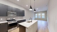 Located at 51 Okner Pkwy in Livingston, NJ, this well-appointed one-bedroom, one-bathroom residence offers a thoughtfully designed layout with modern finishes throughout. The open-concept kitchen and living area serves as the heart of the home, showcasing dark navy cabinetry with gold hardware, a neutral tile backsplash, and a full suite of stainless steel appliances including a gas range, microwave, and French door refrigerator. A central kitchen island with a white quartz countertop and undermount sink provides additional prep space, while two sleek pendant lights overhead add a contemporary touch. Light-toned hardwood flooring flows seamlessly throughout the entire residence. The living area benefits from a bank of three large windows fitted with roller shades, allowing ample natural light and views of the surrounding neighborhood. The spacious primary bedroom mirrors this bright, airy quality with its own set of triple windows and additional single window. The bathroom features marble-look tile flooring and white subway tile surrounding a full tub-shower combination. Additional conveniences include a dedicated laundry area, a utility room, and three closets, providing generous storage throughout the residence.