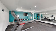 Located at 3225 Long Beach Blvd in Long Beach, CA, this fitness center is a well-appointed, single-room exercise facility designed to support a well-rounded workout routine. The space is finished with durable rubber-matted flooring and clean white ceilings with recessed lighting, creating a bright and functional atmosphere.

A bold teal accent wall energizes the main workout area, which is equipped with a multi-function cable weight machine, a treadmill, a spin bike, and a rack of colorful medicine balls. A wall-mounted flat-screen television adds an entertainment option during cardio sessions. Full-length mirrors line one wall, enhancing the sense of space and allowing users to monitor their form.

Natural light fills the room through a set of French doors and flanking sidelights, which open directly onto an outdoor pool area — offering a seamless connection between indoor exercise and outdoor amenities. The pool, lounge chairs, and surrounding greenery are visible through the glass, adding a refreshing outdoor ambiance to the interior.

The layout is efficiently organized to maximize usability within its compact footprint, making it a practical and inviting fitness amenity for residents or guests of the property.