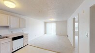 Located at 2721 Montavista Pt, Unit 213 in Springfield, Illinois, this single-floor residence features a clean, neutral design throughout. The open-concept living and dining area is generously sized, finished with soft wall-to-wall carpeting and a textured ceiling. Natural light fills the space through a sliding glass door fitted with vertical blinds, which provides both brightness and easy access to the outdoors. The kitchen is efficiently laid out with light wood cabinetry extending to the ceiling for ample storage, a full-size refrigerator, electric range, dishwasher, and a double sink set into a continuous countertop. The tile flooring in the kitchen offers a clean contrast to the carpeted living areas. The bedroom is a comfortable, well-proportioned room with plush carpeting, a closet, and direct sightlines to the kitchen area. The bathroom is thoughtfully equipped with accessibility features, including installed grab bars near the toilet, and is finished with patterned vinyl tile flooring. The overall palette of whites and warm neutrals creates a bright, airy atmosphere throughout the unit, making it a practical and welcoming space.