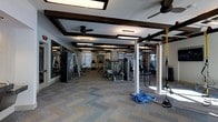 This expansive fitness center is designed to accommodate a comprehensive workout experience across a single, open floor plan. The space features a refined architectural aesthetic, with a coffered ceiling accented by dark wood beams, recessed lighting panels, and ceiling fans that enhance both the visual appeal and ventilation of the room. Large windows allow natural light to fill the space, contributing to an open and energizing atmosphere. The flooring consists of patterned carpet tiles that define the workout areas while adding a polished, modern look. The strength training section is well-appointed with a variety of cable machines, weight benches, and resistance equipment arranged for ease of use and flow. A functional training zone includes a multi-station rig equipped with TRX suspension trainers and battle ropes, ideal for dynamic and cross-training workouts. The cardio section lines one side of the facility with a row of treadmills and other aerobic machines. Full-length mirrors are strategically placed along the walls to maximize the sense of space and support proper exercise form. The overall layout is thoughtfully organized to separate training zones while maintaining an open, unobstructed feel throughout the facility.
